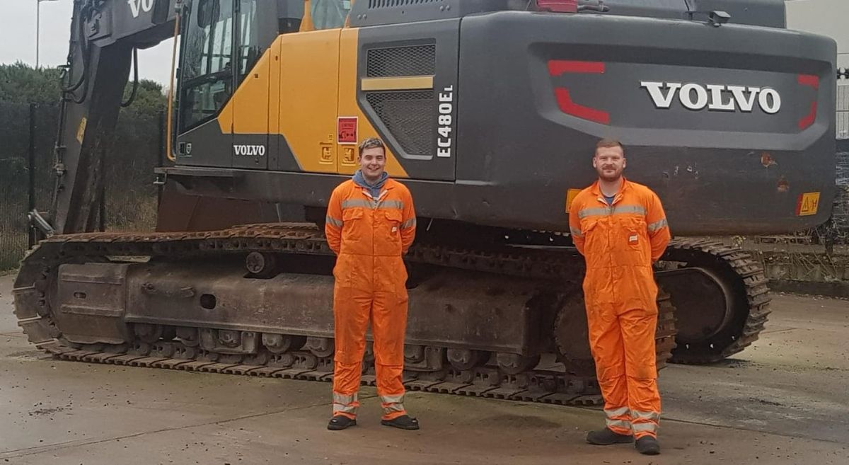 Two men in hi viz work suits standing in front of a Volvo excavator.